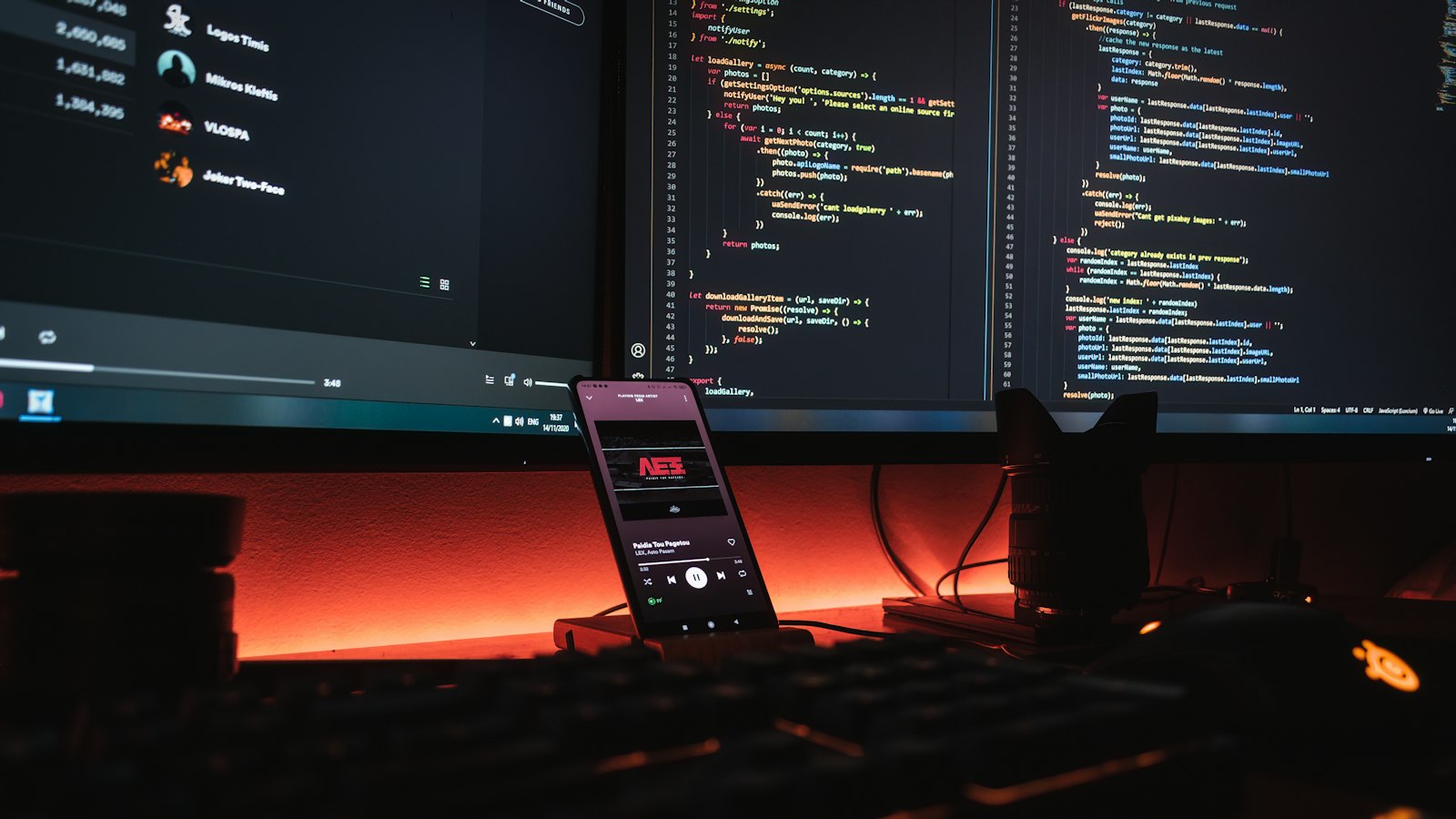A smartphone displaying music on a desk with computer monitors showing code